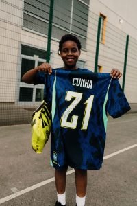 A smiling young player holding up a signed Cunha number 21 Brasil shirt with his JD Sports gift bag