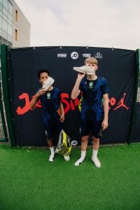Two young players in blue Brasil kits holding up their new Jordan trainers in front of the Joga Sinistro backdrop