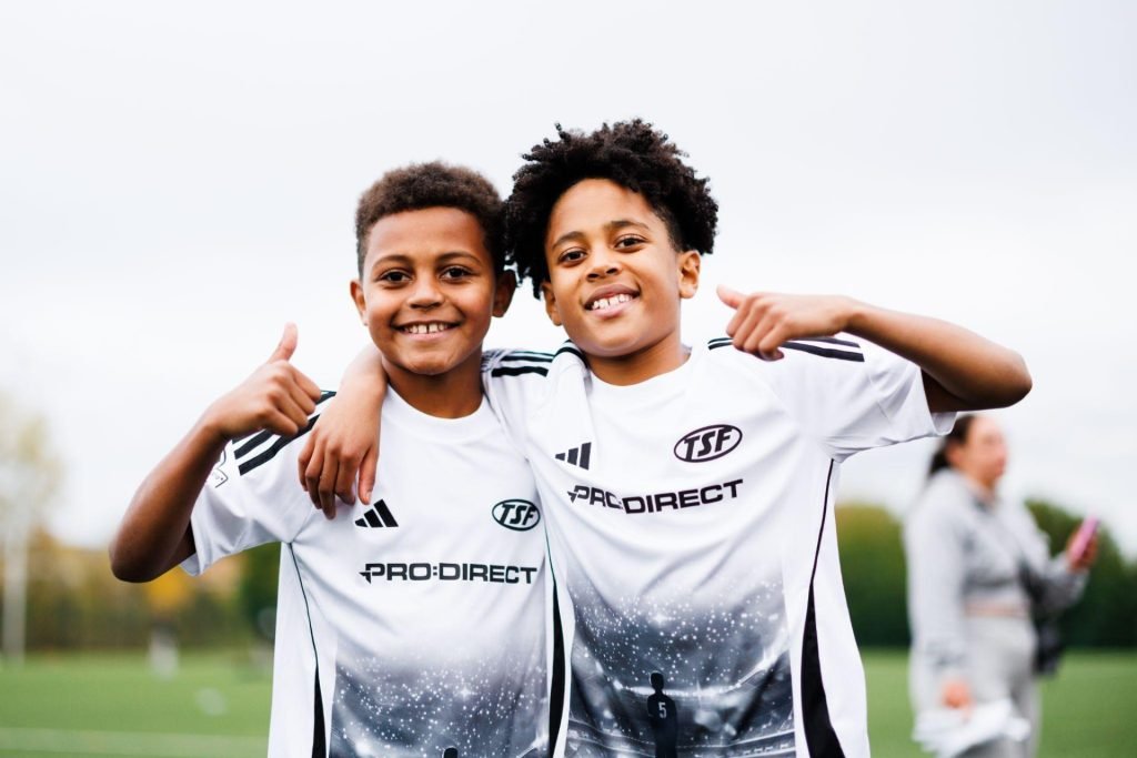 Two young TSF players in Pro:Direct and adidas kits smiling together with their arms around each other on the pitch