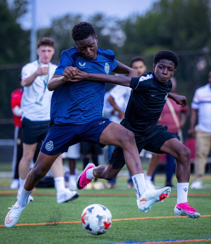 Two young players in blue and black kits battling shoulder to shoulder for the ball on the pitch