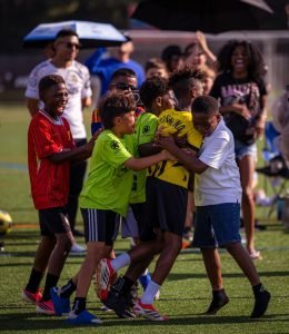 Young players in bright kits mobbing a teammate in a yellow Dortmund shirt to celebrate a 1v1 win
