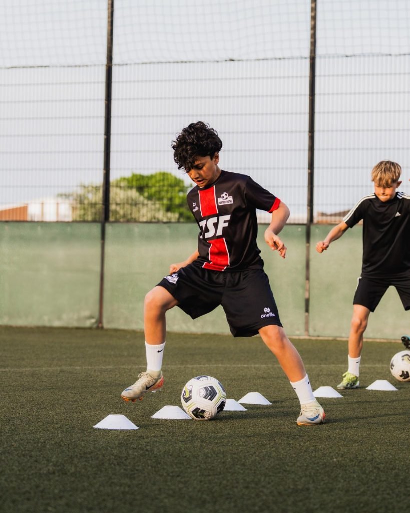 Young TSF player in a red and black kit dribbling through cones at academy training