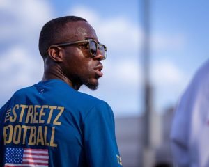 A TSF coach photographed from behind, his blue shirt showing The Streetz Football lettering in gold beside a USA flag