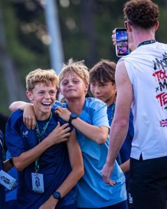 Two young players hugging and laughing while a TSF team member films them on a phone at the Rising Stars Cup