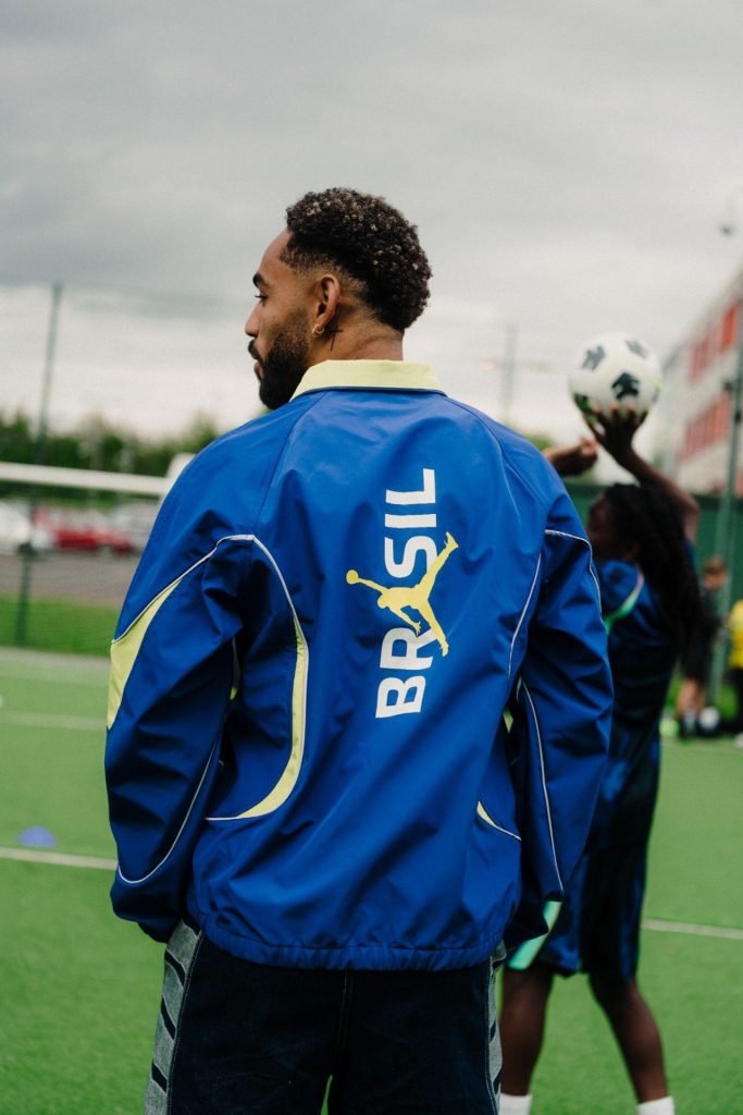 Matheus Cunha in a blue Jordan Brasil jacket looking back over his shoulder as a young player lifts a ball behind him