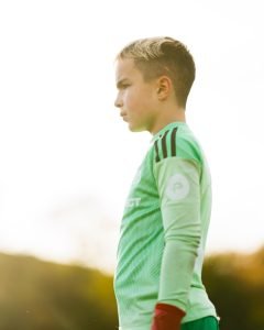 A young TSF goalkeeper in a green kit in profile against bright evening sunlight