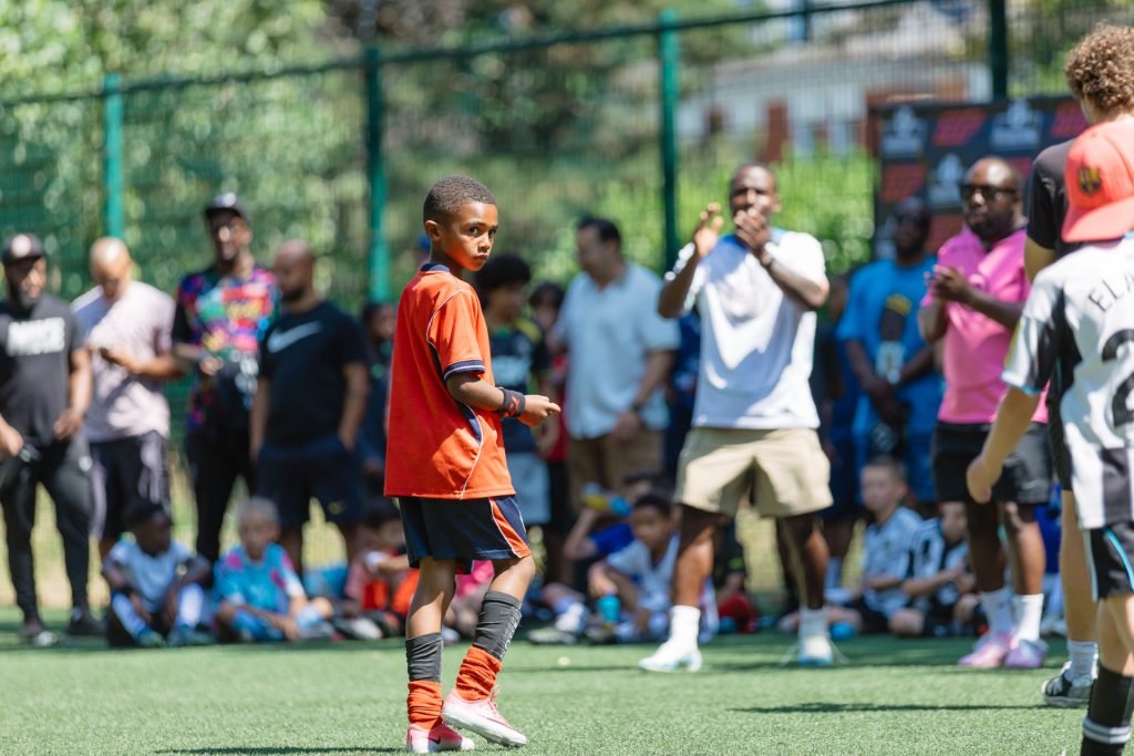 A young player in an orange kit facing up his opponent as a crowd of parents cheers pitchside