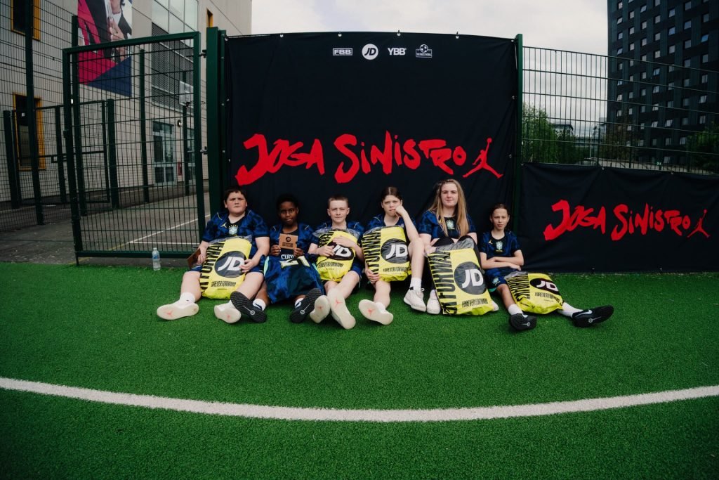 Six young players in blue Brasil kits sitting on the pitch with their yellow JD Sports bags in front of the Joga Sinistro backdrop
