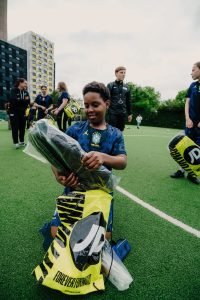 A young player kneeling on the pitch unwrapping new boots from his yellow JD Sports bag
