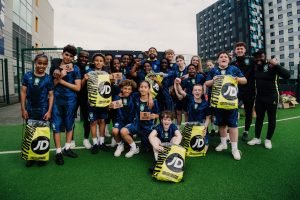 Young players and coaches celebrating together on the pitch, holding up JD Sports bags and gift cards